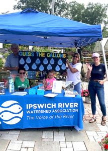 A group of volunteers are standing outdoors under a blue tent, holding a sign that says guess the gallons. There is a table with a blue tablecloth and the IRWA logo.